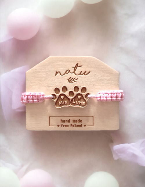 Bransoletka z drewnianą zawieszką przedstawiającą dwie łapki psa z grawerem imion piesków, na różowym, ręcznie plecionym sznurku.
